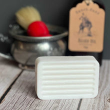 Bar of soap on a wooden surface with a brush and container in the background.