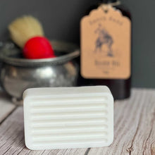 Bar of soap on a wooden surface with a blurred background featuring a bottle and brush.