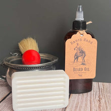 Beard oil bottle with a label, brush, and soap on a wooden surface.