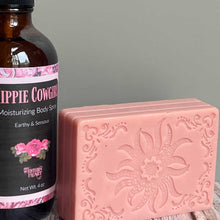 Bottle of 'Hippie Cowgirl' moisturizing body spray next to a pink soap bar with floral design on a gray background
